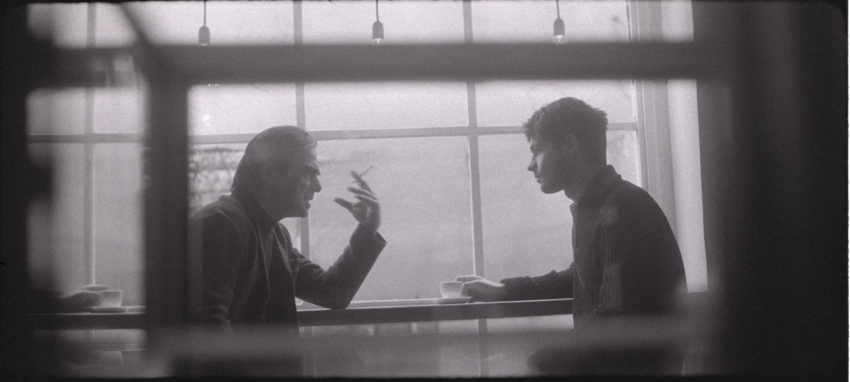 Two men talk over coffee at a café window, seen through the glass.