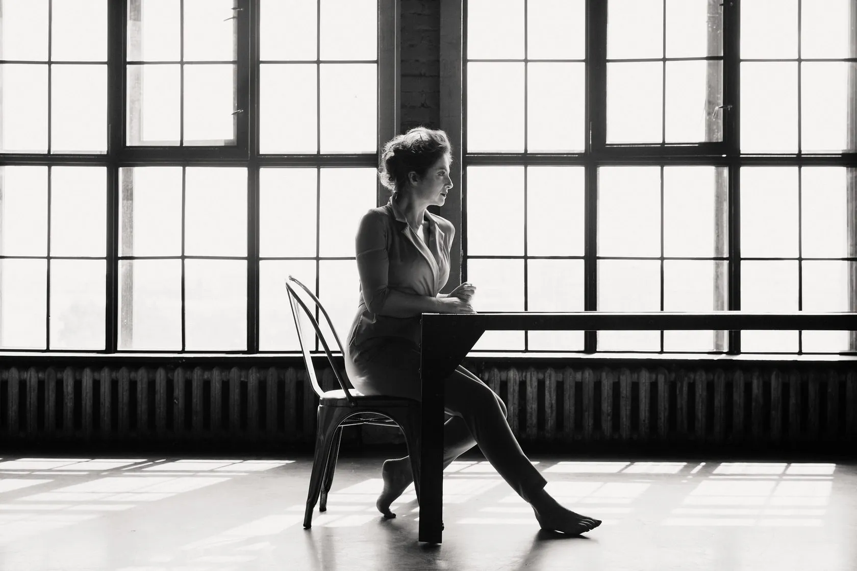 Anna Banshchikova sits at a table by a tall factory window, in black and white.