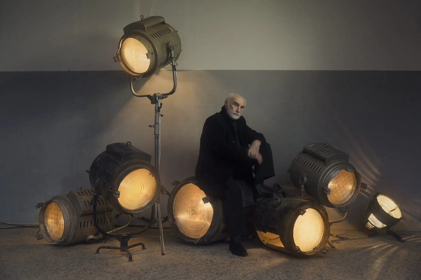 A white-bearded man in black sits among old glowing studio spotlights.