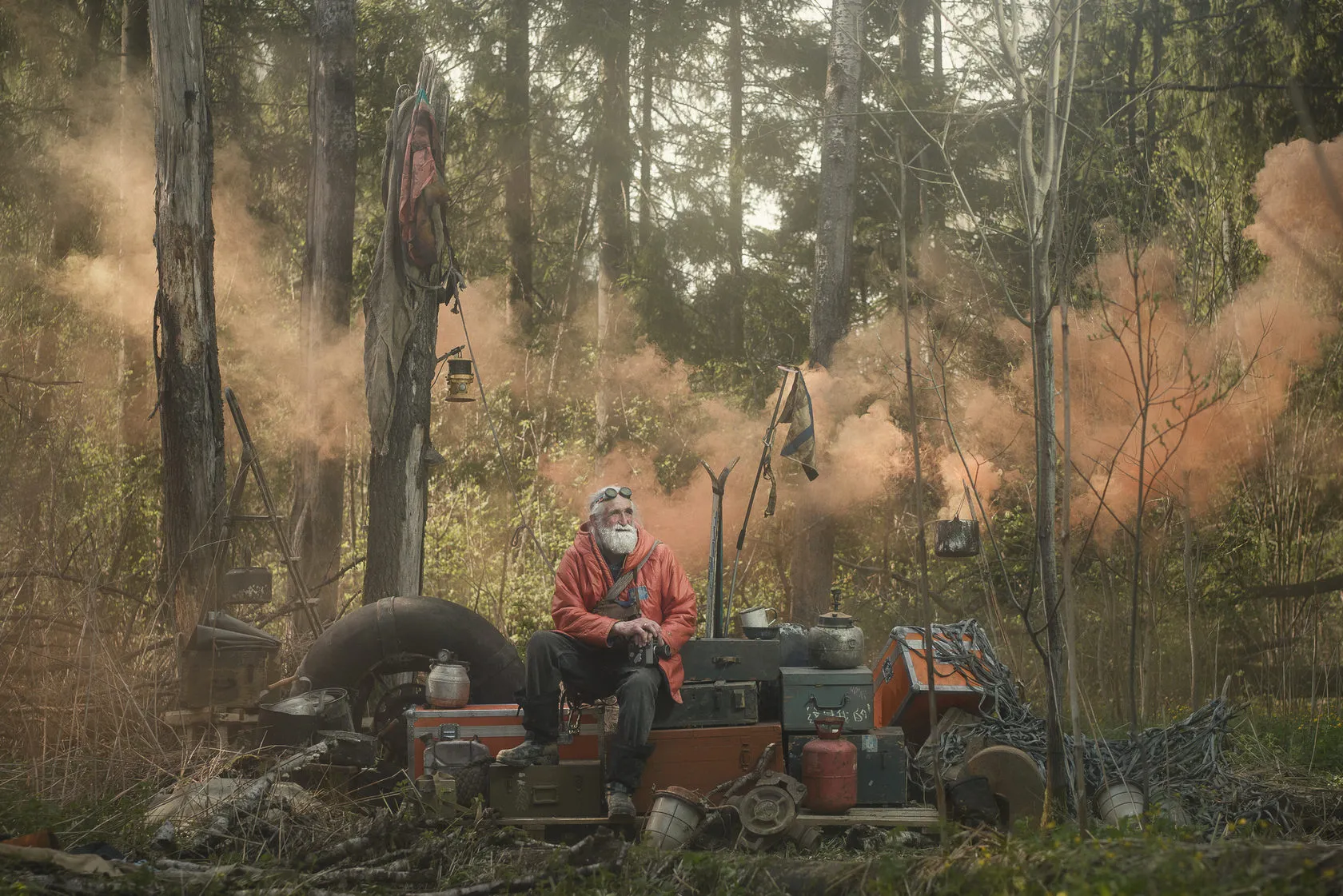 Vladimir Chukov in a red parka sits on old luggage in a forest clearing as orange smoke drifts through the trees.
