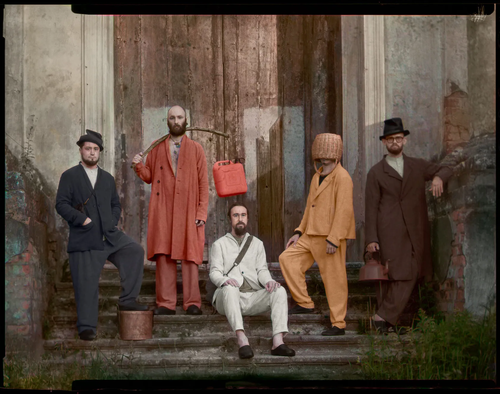 Five men in loose Infundibulum suits and hats pose on the steps of a ruined building.
