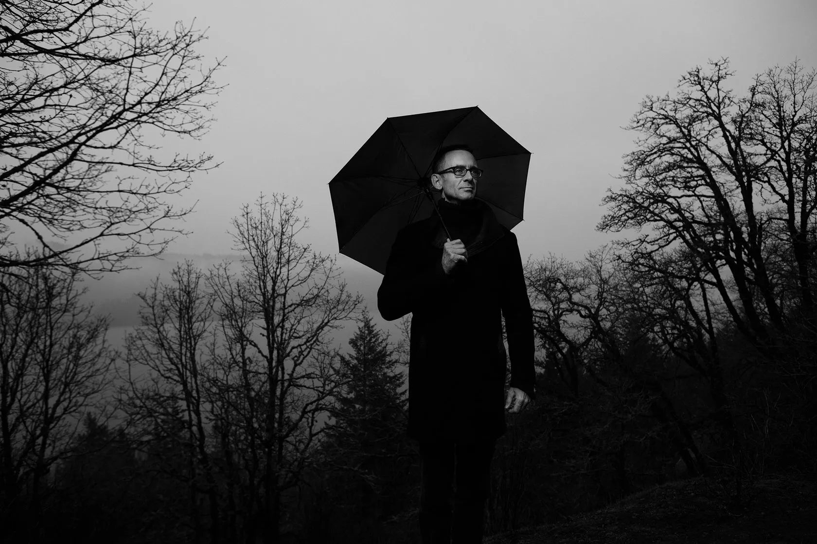 Chuck Palahniuk stands under a black umbrella among bare trees.
