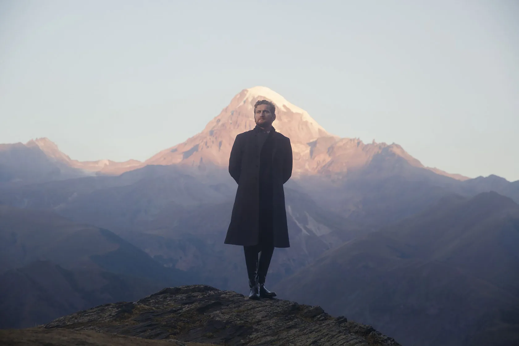 Jimsher Chkaidze in a long black coat stands on a ridge before a snow-capped peak.