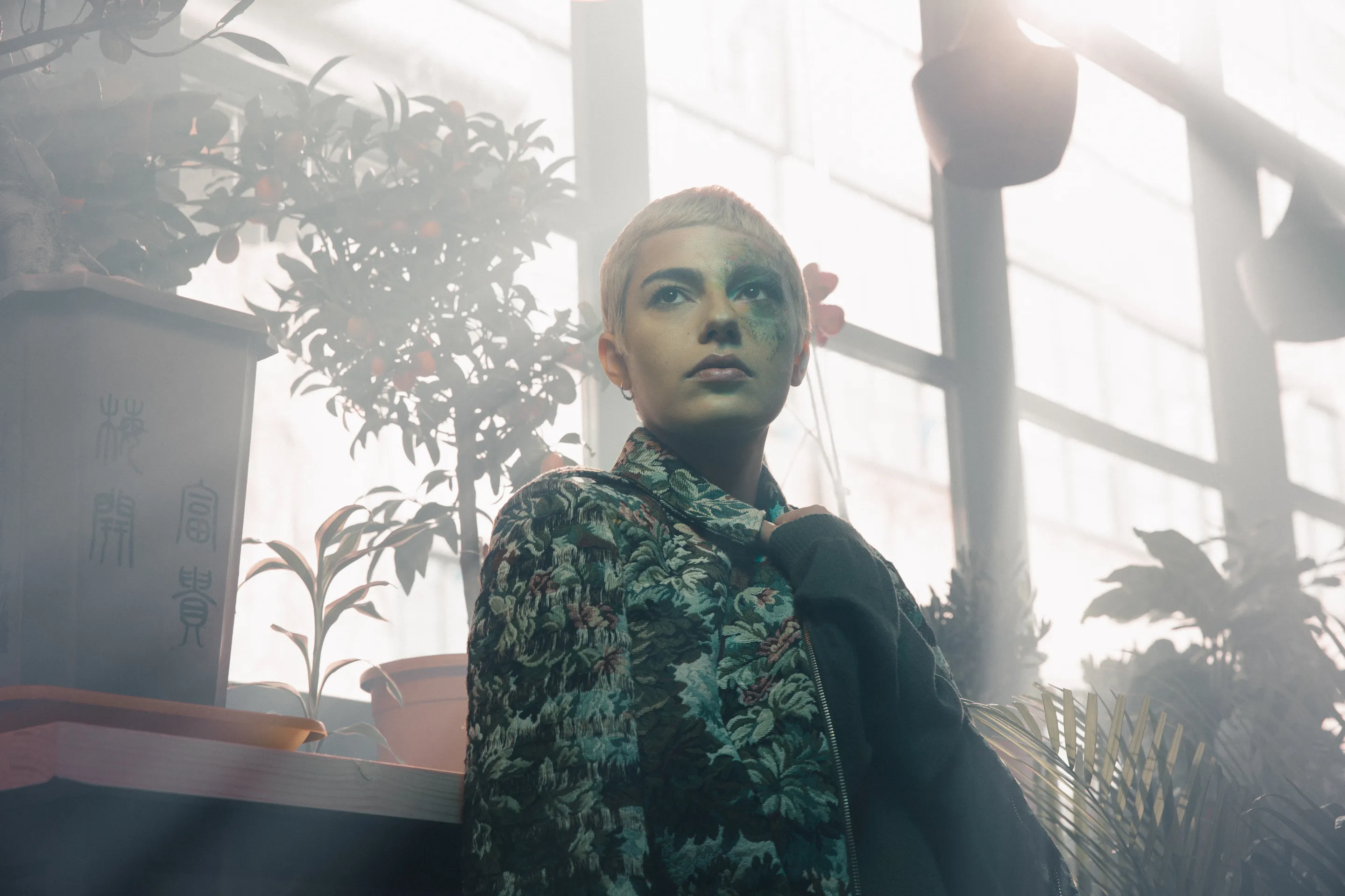 The Jacket campaign by Andrei Kovalev: a woman with pale makeup stands by a sunlit window among houseplants.