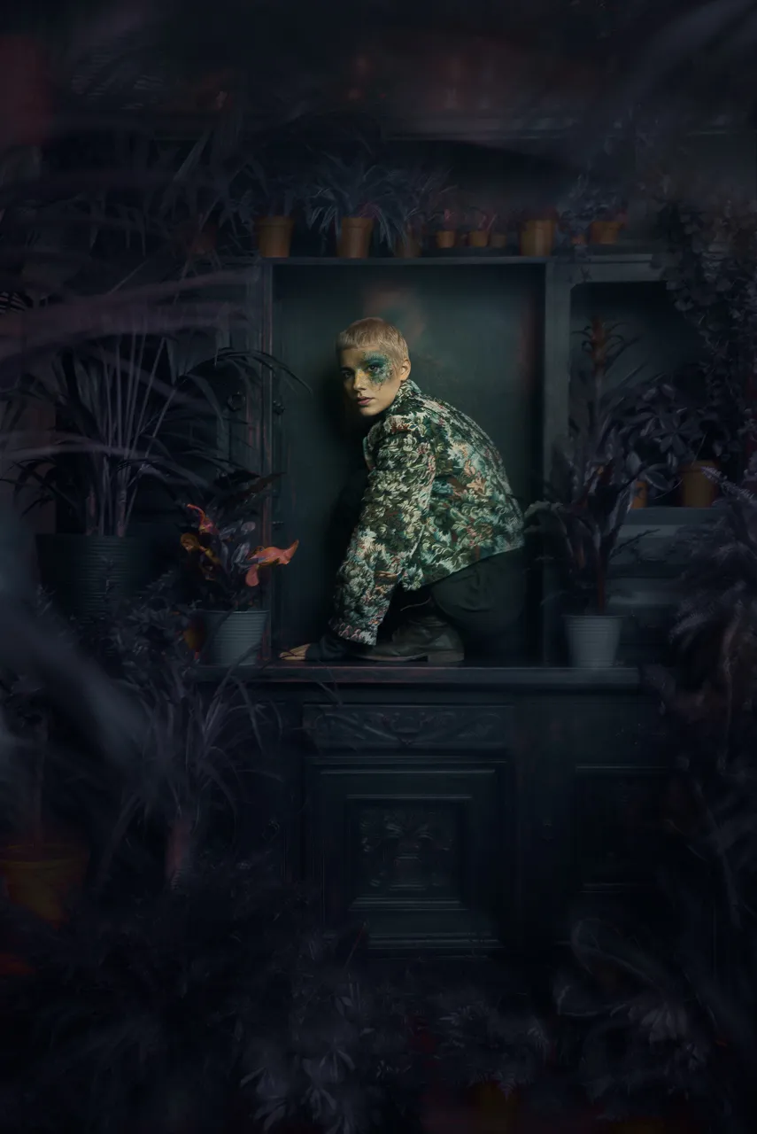 The model crouches in a dark wooden niche among houseplants, in a floral jacket.