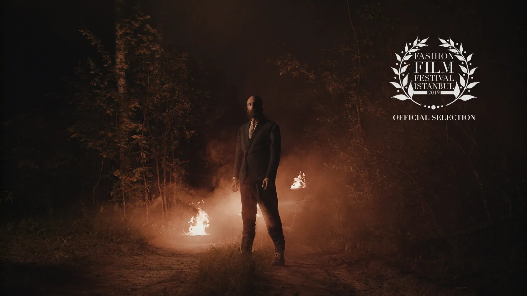 A bearded man in a suit stands on a forest path at night between small fires.