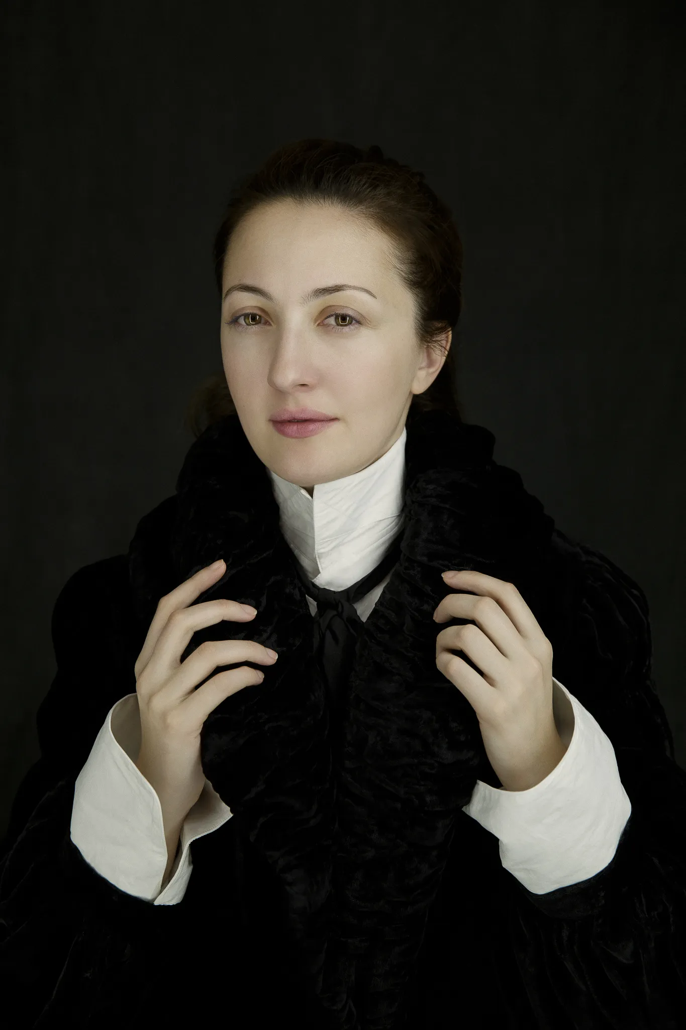 A woman in a black velvet coat and high white collar holds its lapels.