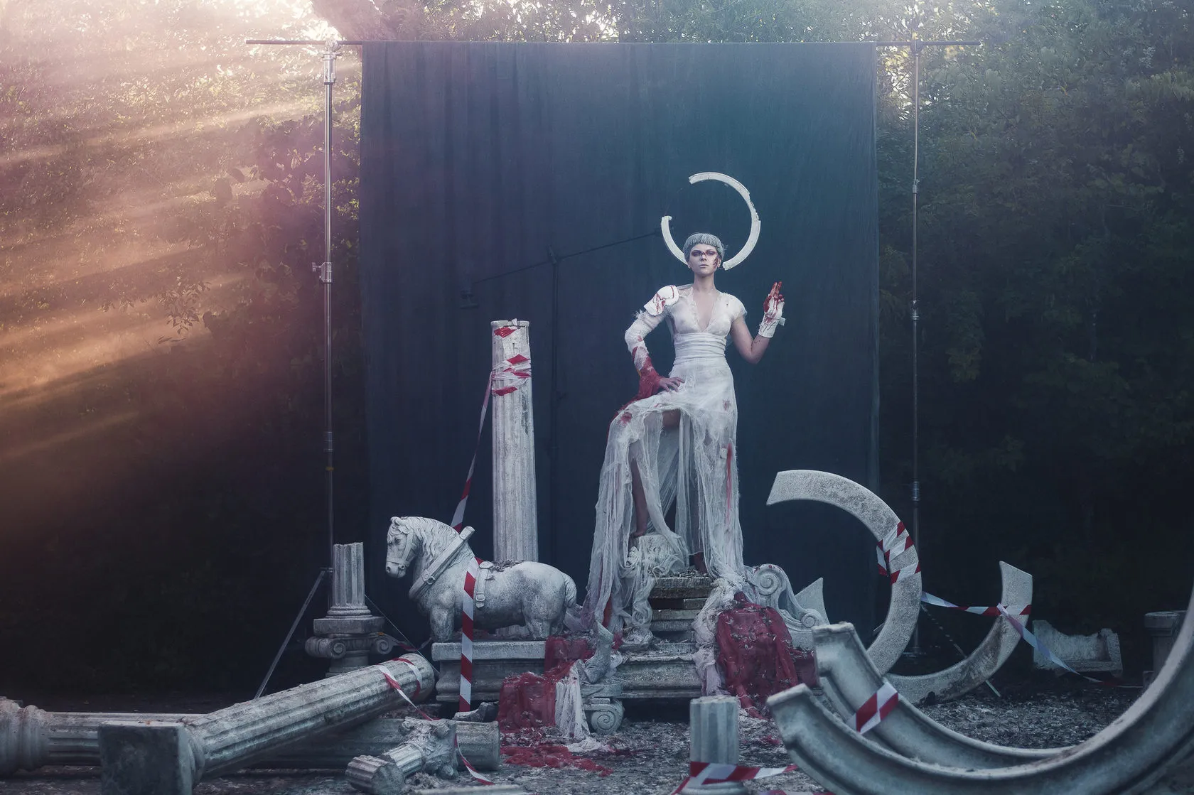 A woman in a torn white gown and a broken halo stands among fallen columns and a stone horse.