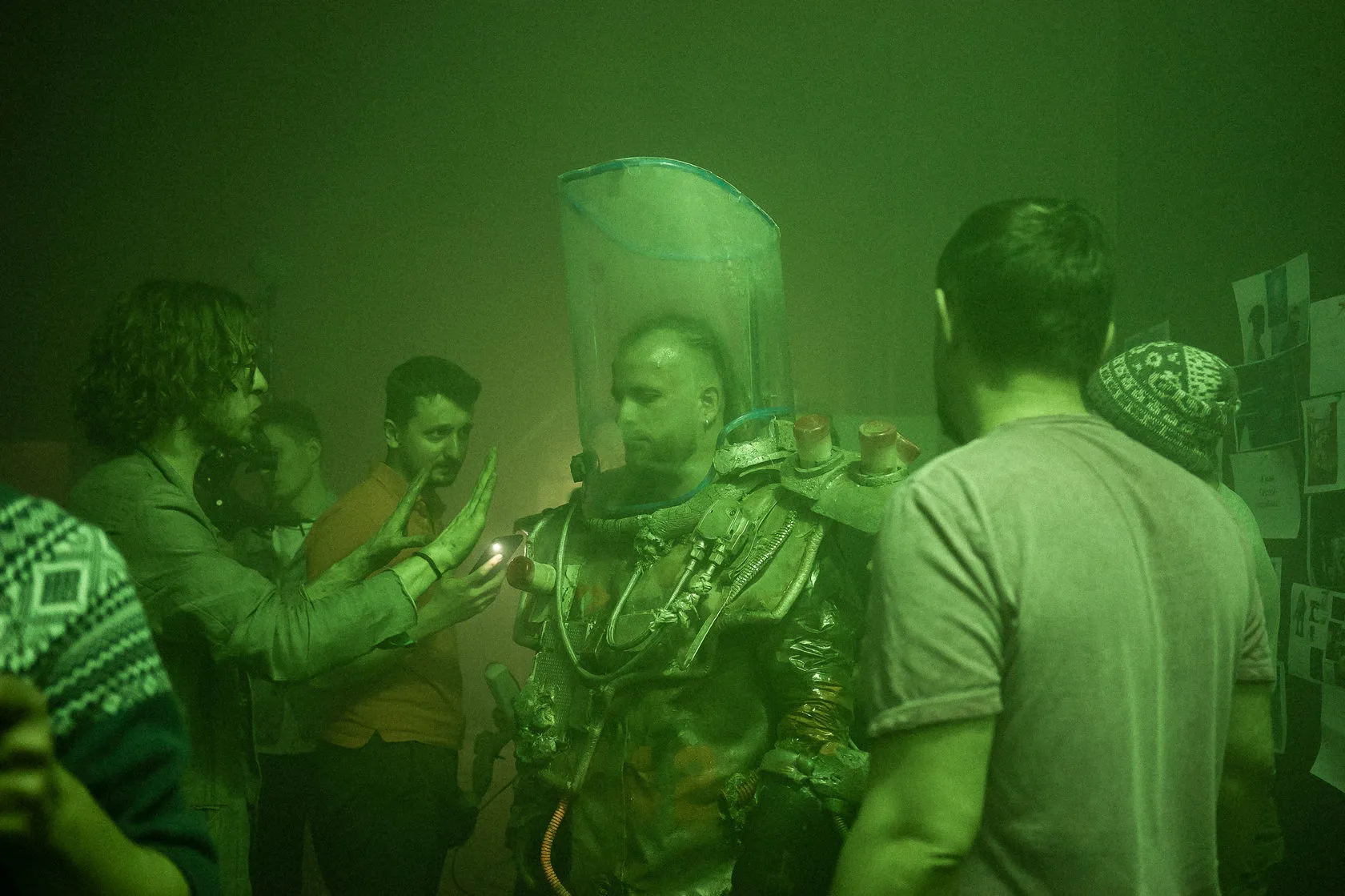 A crew gathers round an actor in a rusty suit and tall clear helmet on a green-lit set.