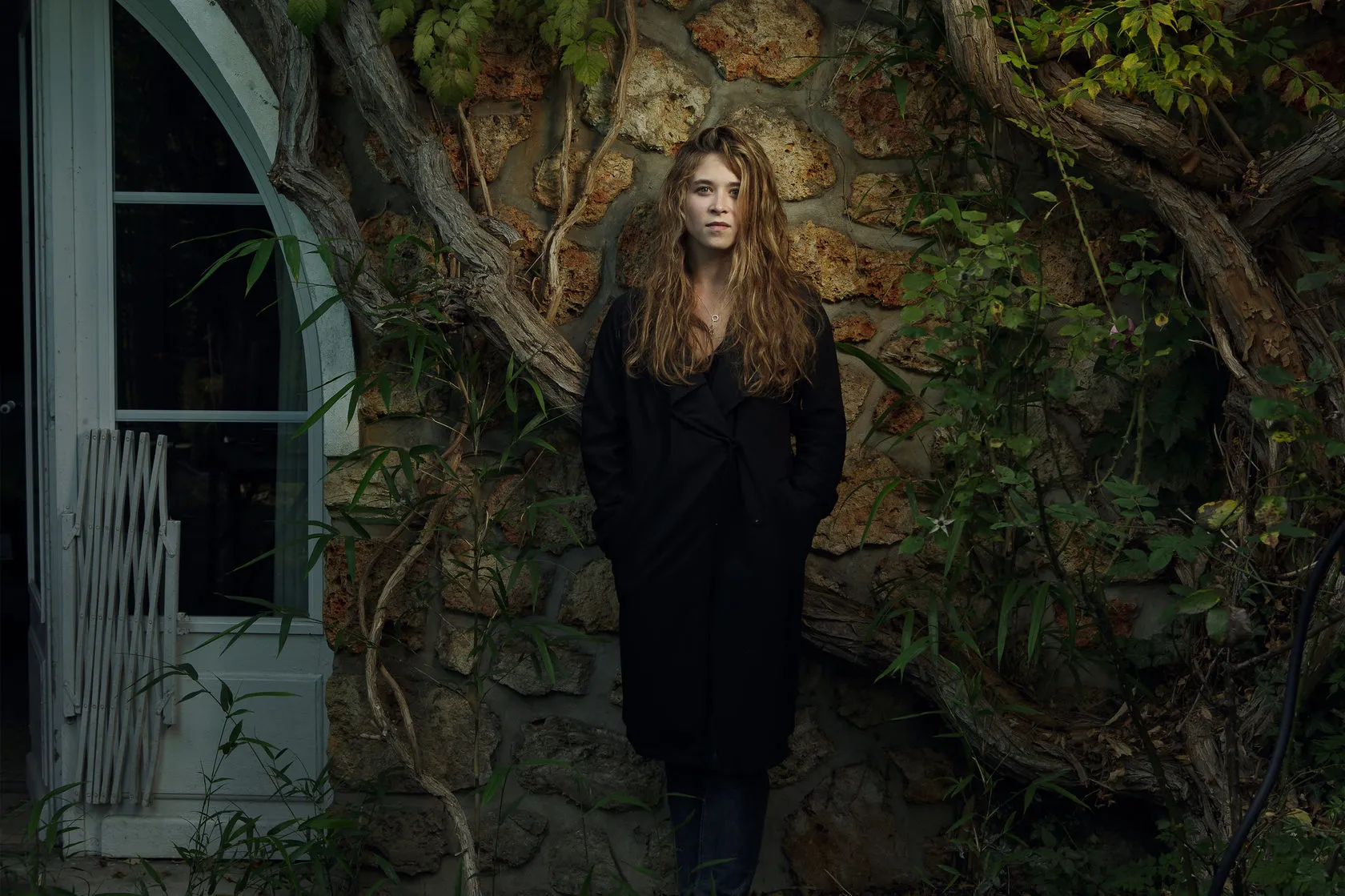 Emma stands in a black coat against a stone wall overgrown with vines.
