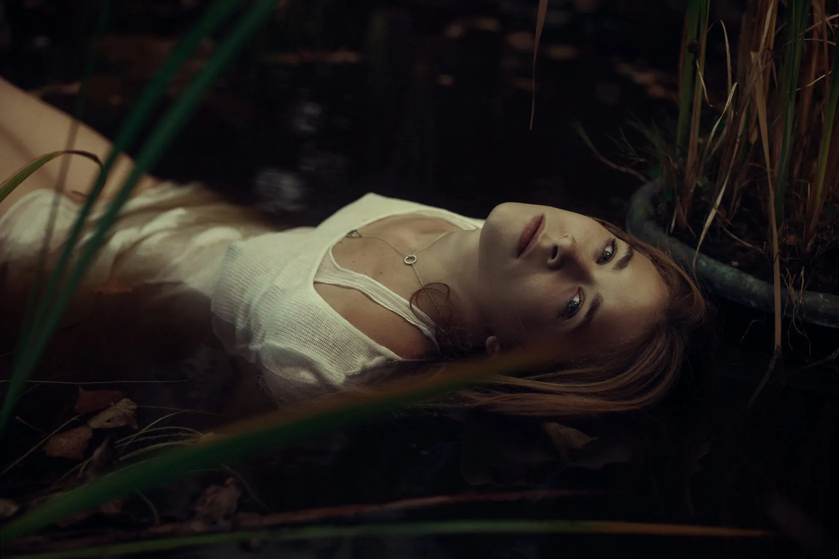 Emma lies in dark water among reeds, eyes open, in a white dress.