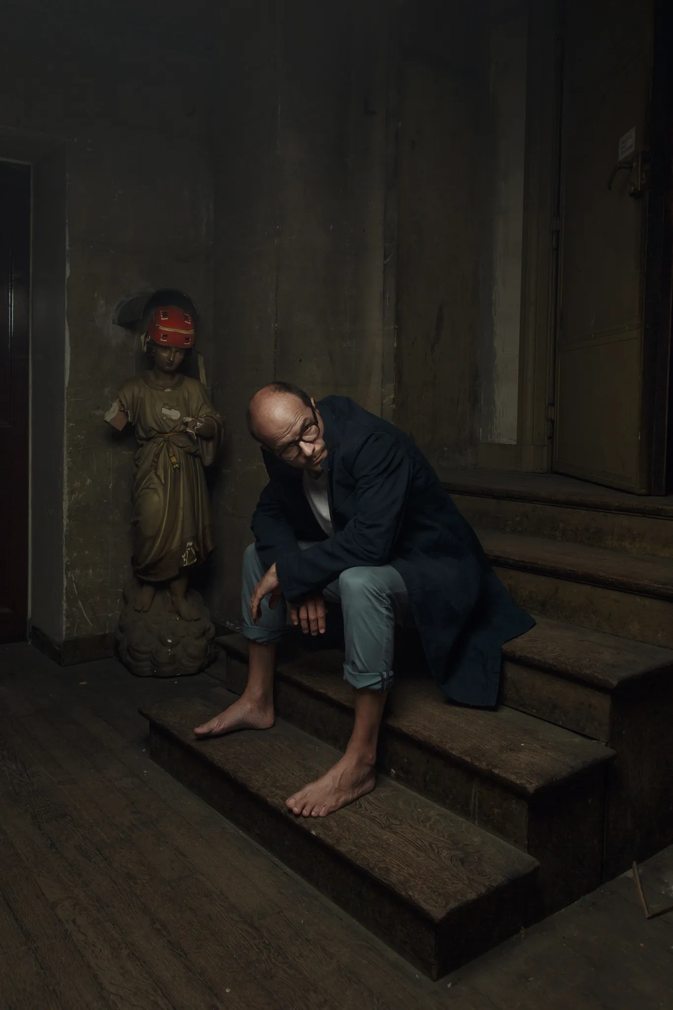 Laurent Clerc sits barefoot on wooden stairs beside a saint’s statue with a red blindfold.