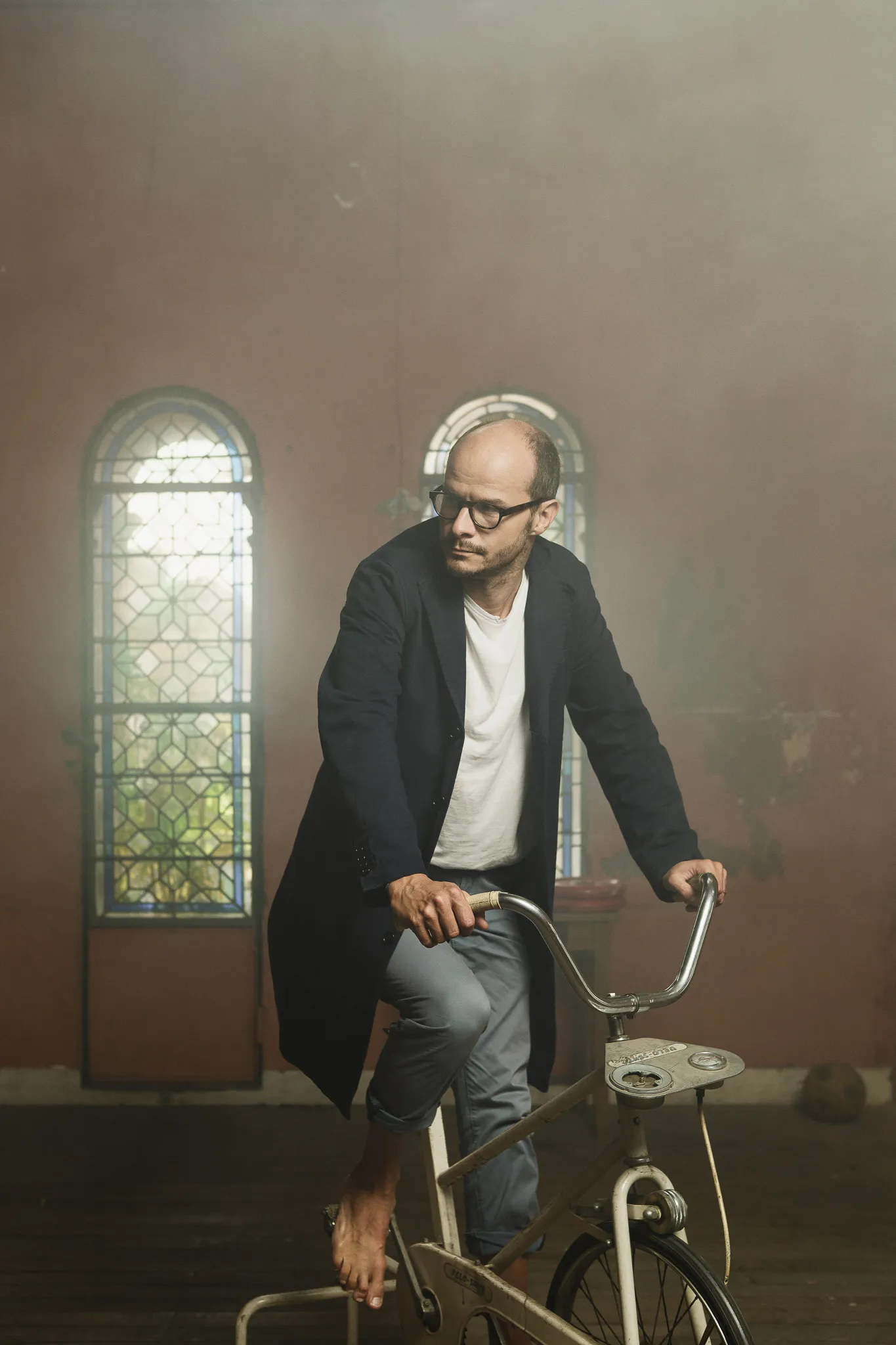 Laurent rides an exercise bike barefoot beside a stained-glass window.