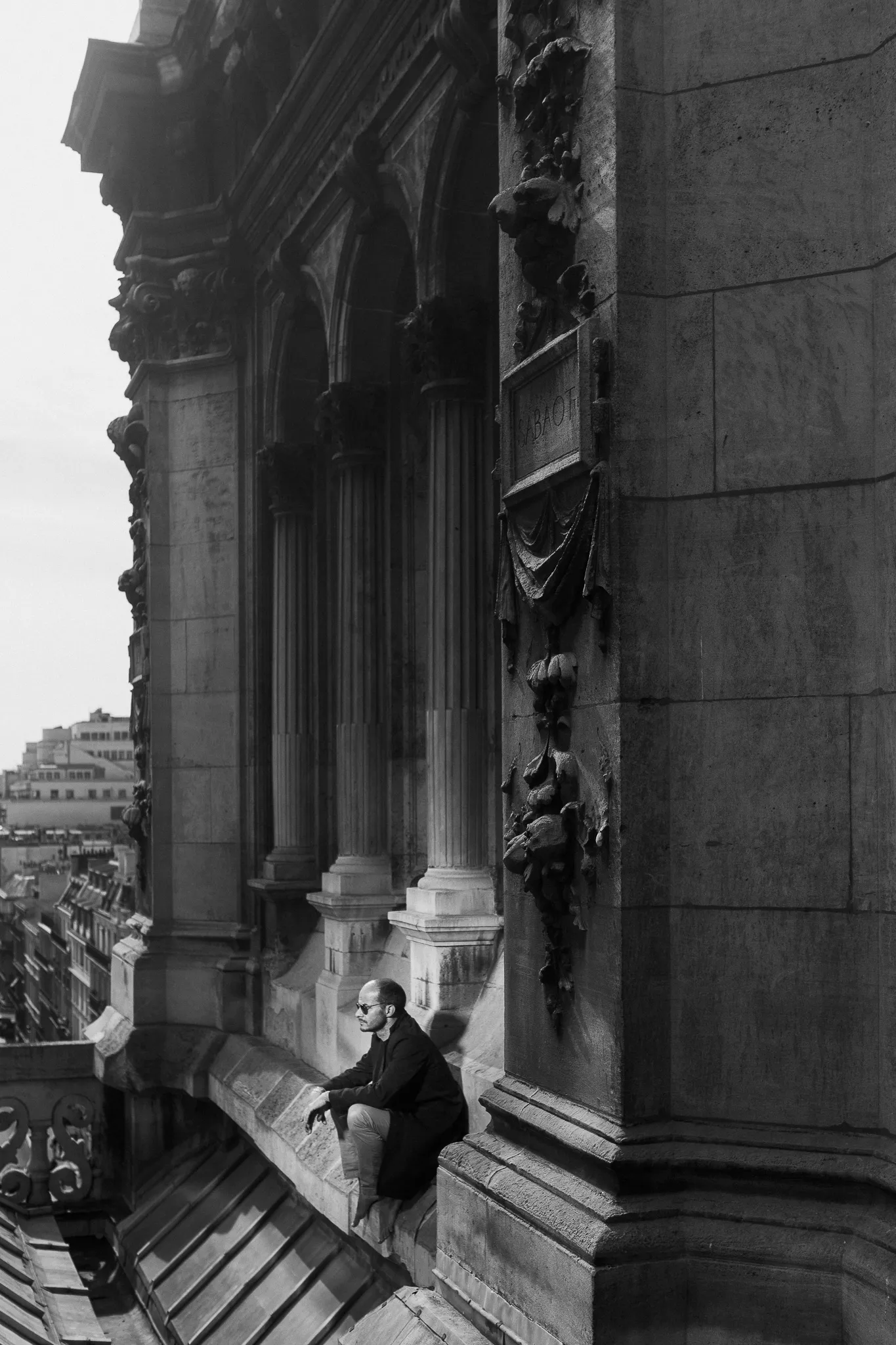 Laurent crouches on a narrow roof ledge below carved stone columns, in black and white.