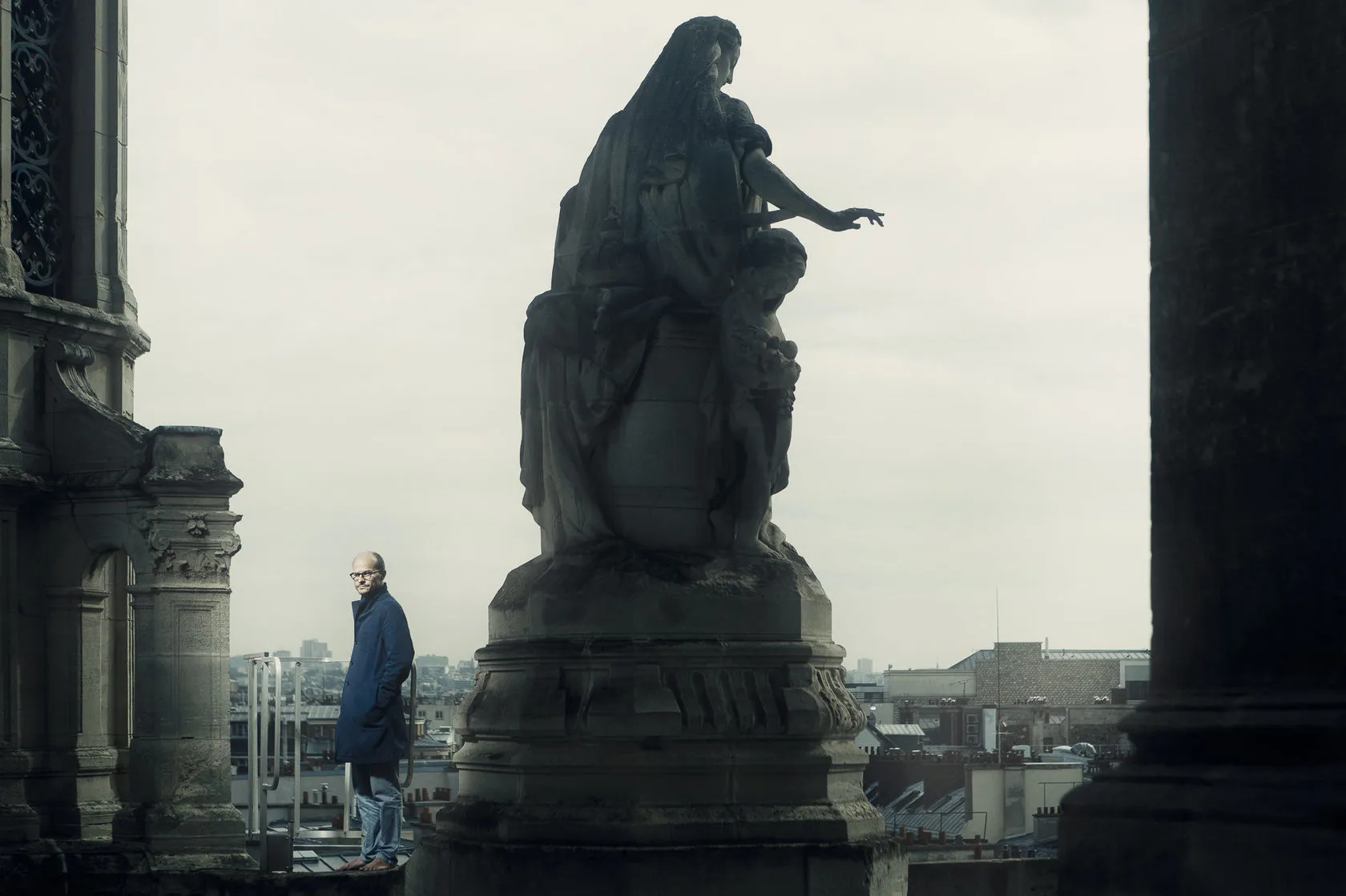Laurent stands on the church roof beside a large stone statue, the city behind.