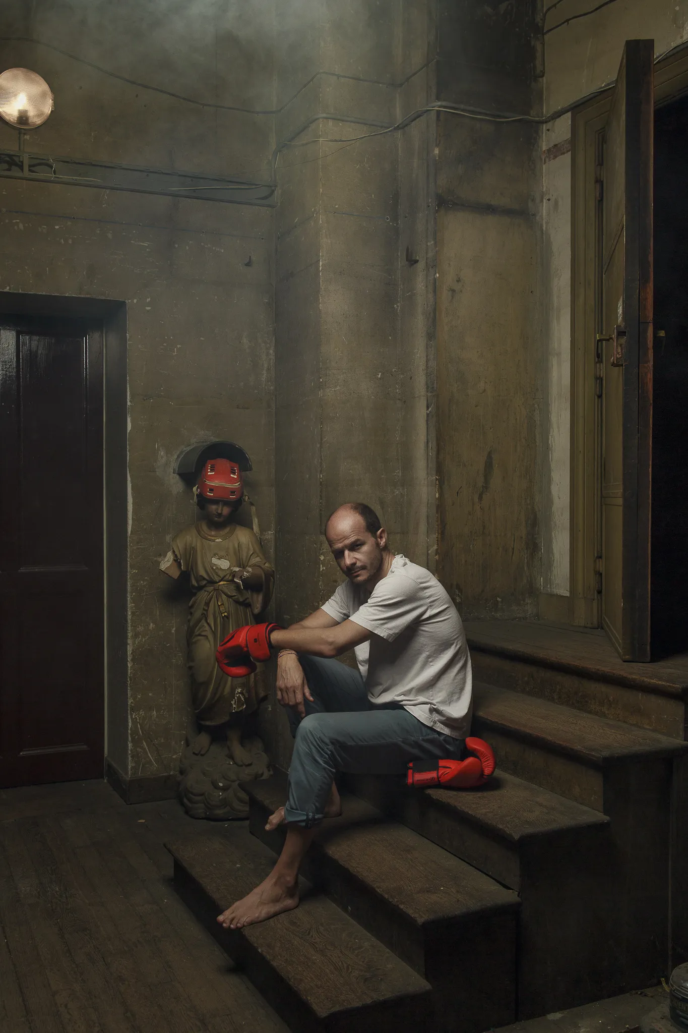 Laurent sits barefoot on the stairs in a white T-shirt, next to the blindfolded statue.