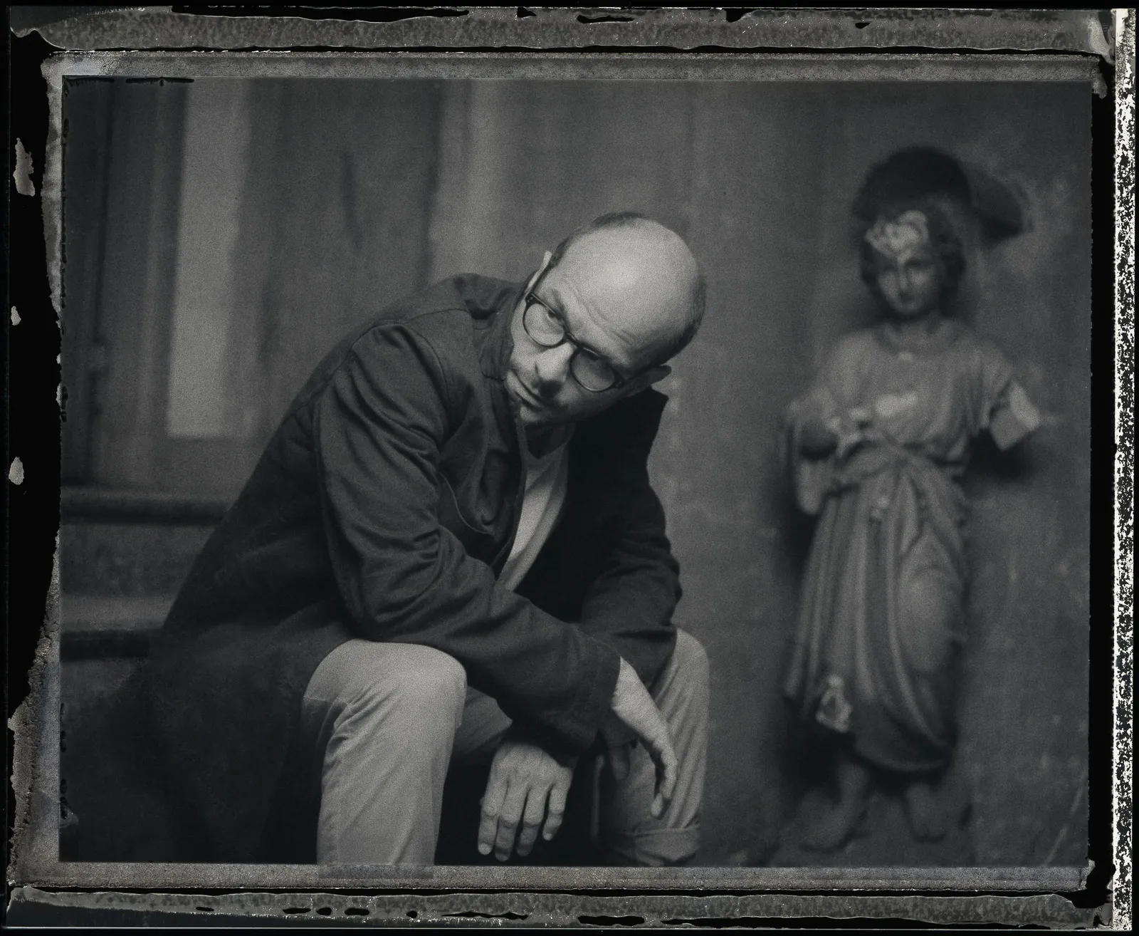 Laurent sits by a statue of a saint, chin in hand, on old instant film.