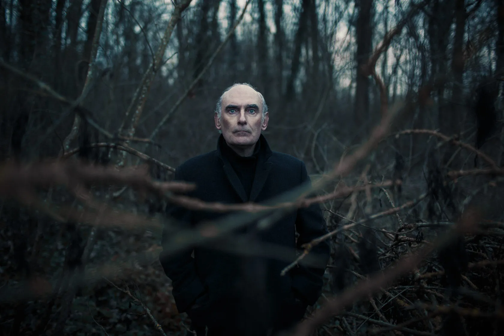 Michel stands among bare branches in a winter wood, looking into the camera.