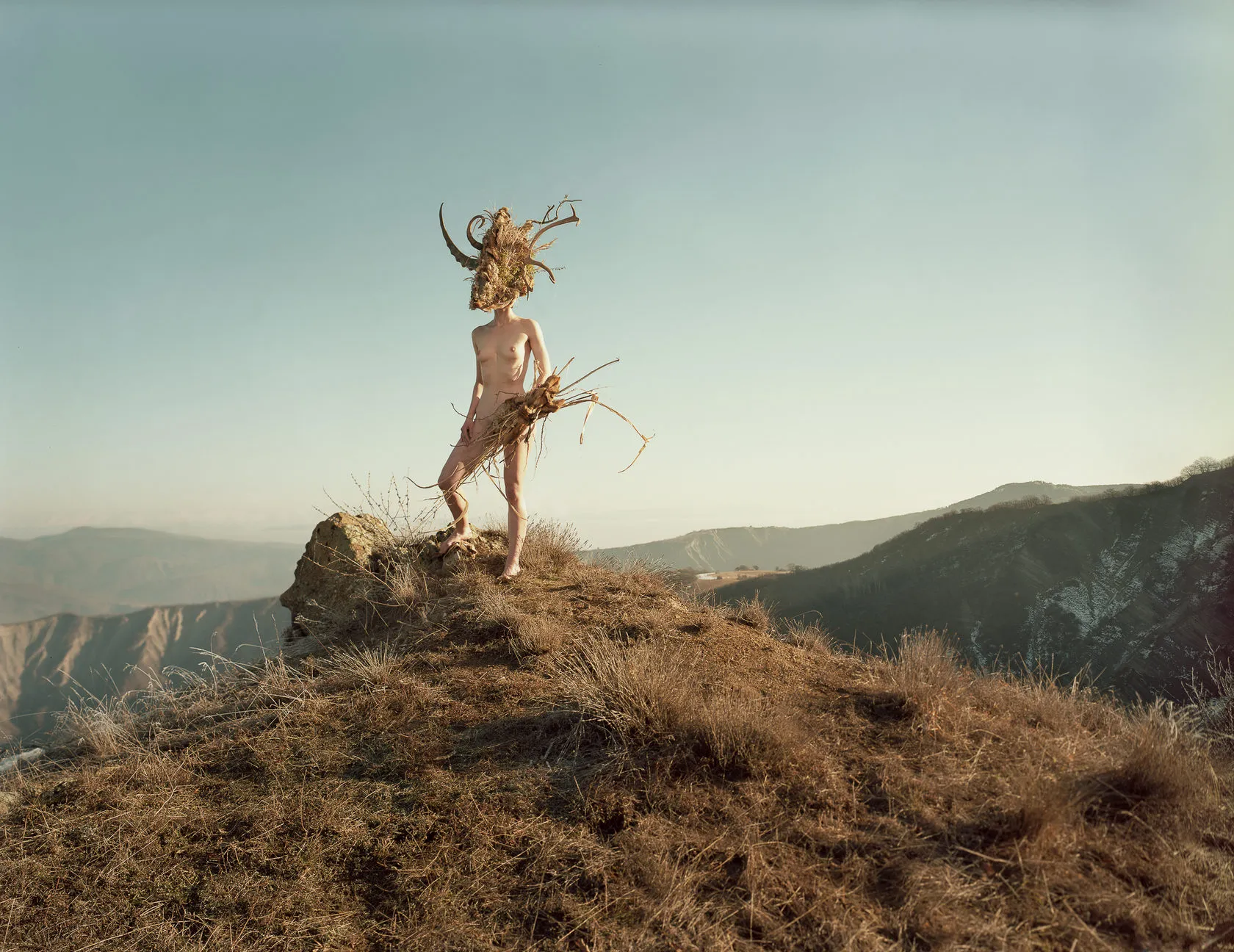 Dàli stands on a rocky outcrop above a mountain valley, holding a bundle of dry grass.