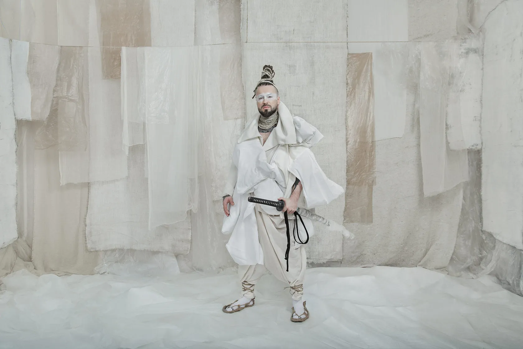 A Burito musician in white robes and a topknot, a sword at his belt, before patchwork white drapes.