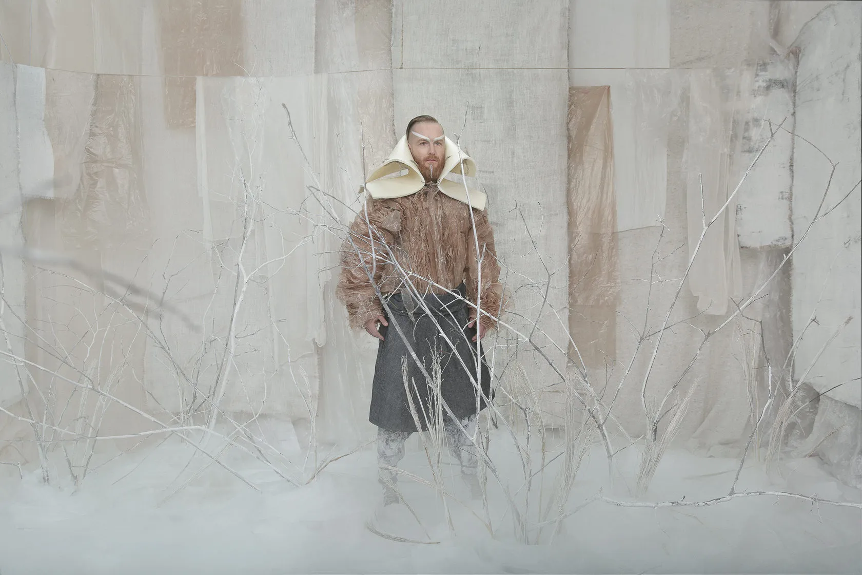 A man in a brown fur coat with a tall white collar stands among bare white branches.