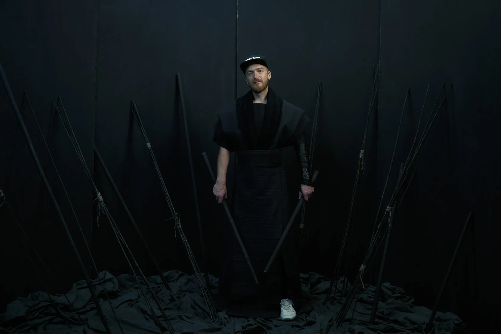 A Burito musician in a cap and black costume among black spears, from the 2015 shoot.