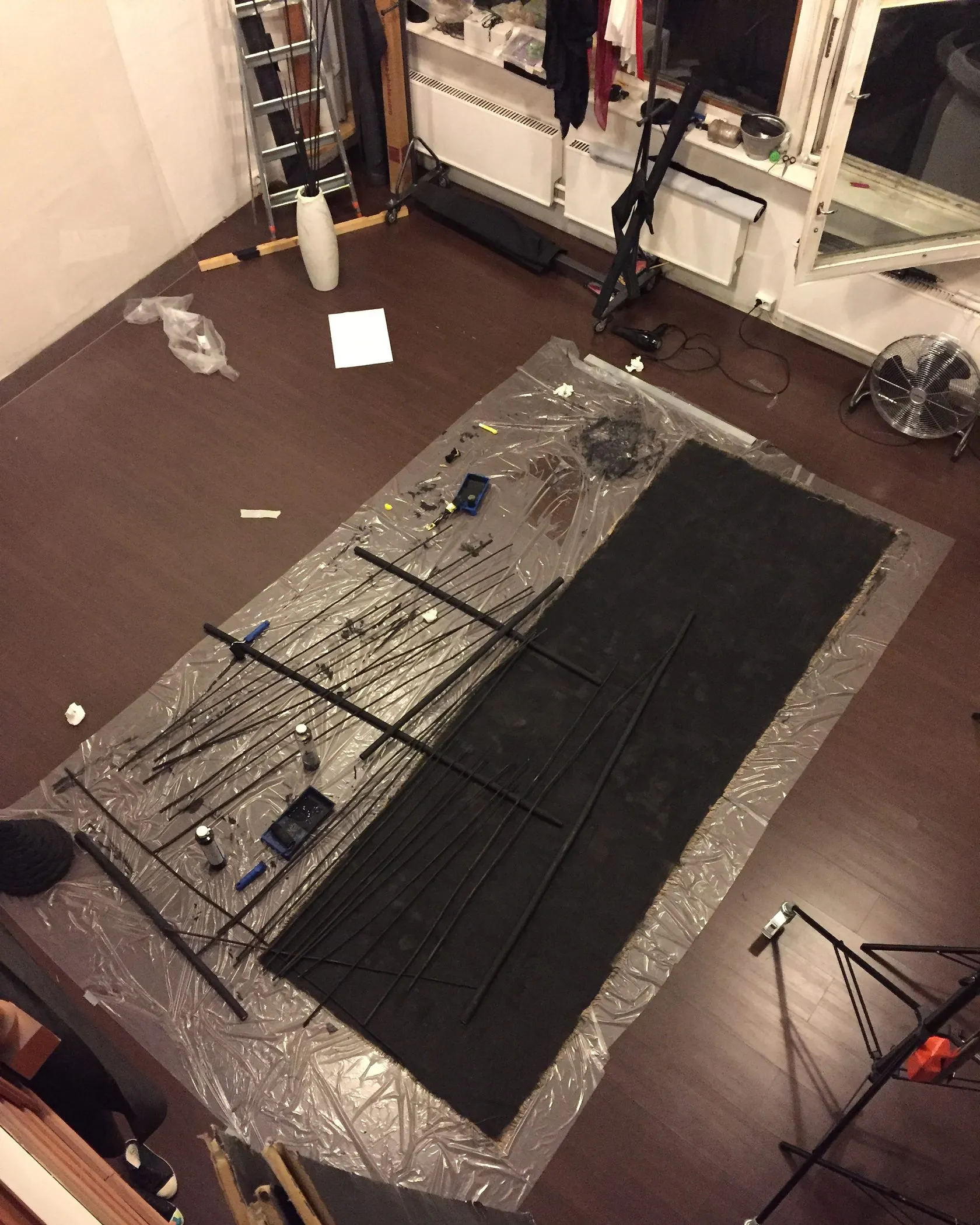 Seen from above, black cloth and poles laid out on plastic sheeting on a studio floor.