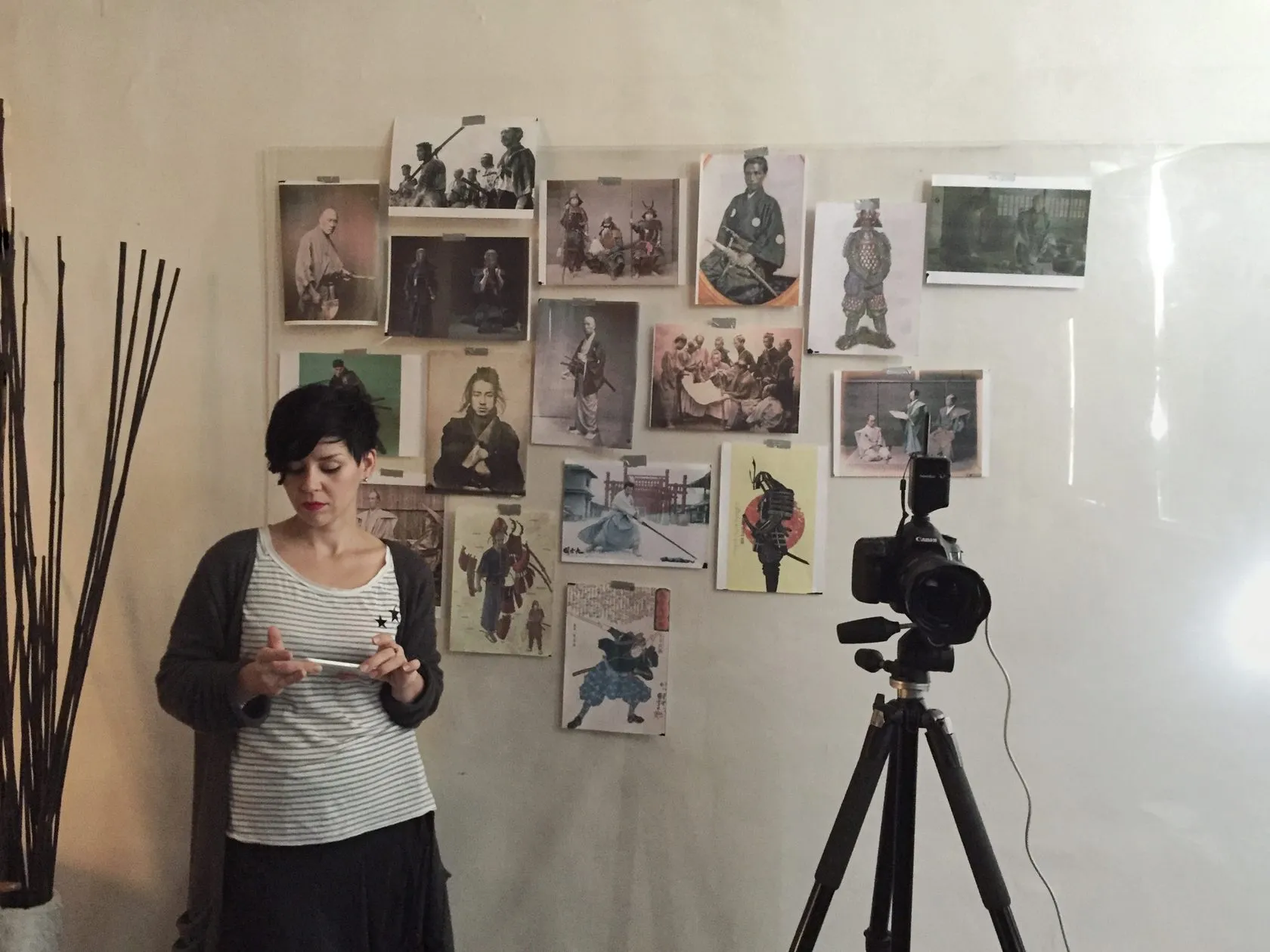A woman checks her phone by a wall of samurai photos and prints, next to a camera on a tripod.