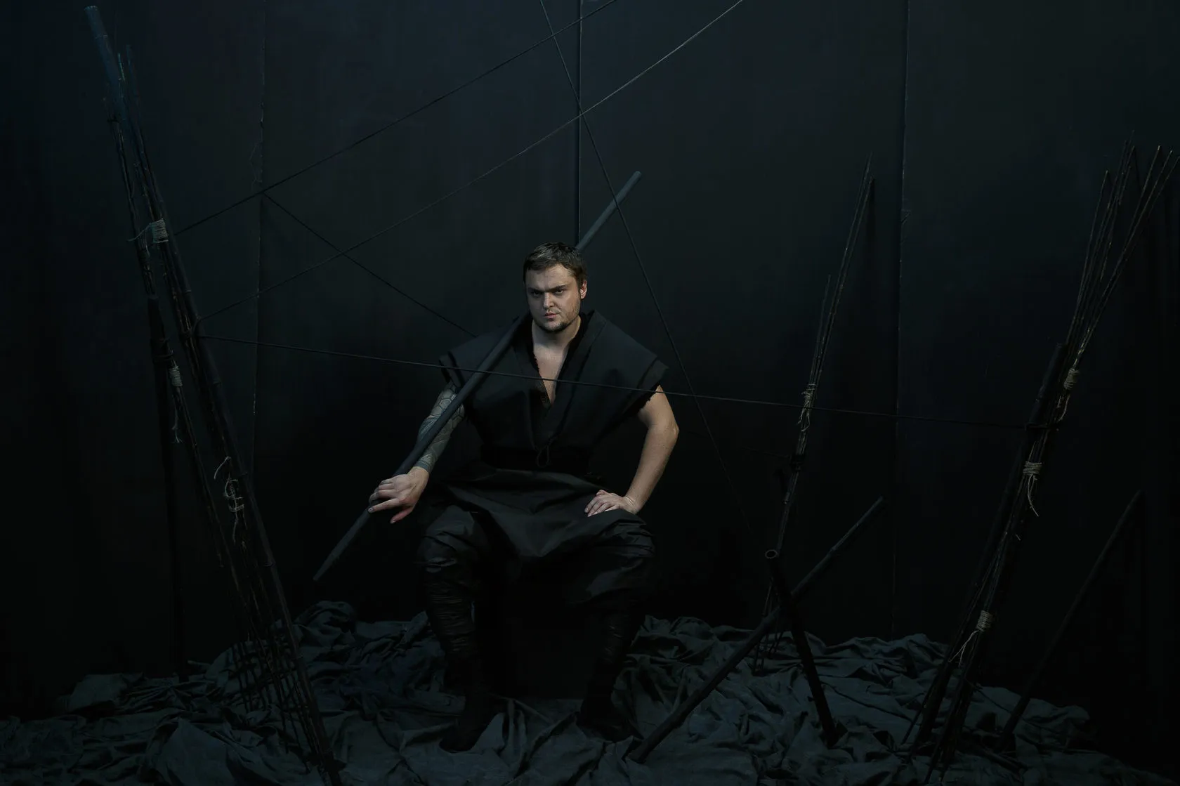 A Burito musician sits with a staff across his knees among tall black poles.