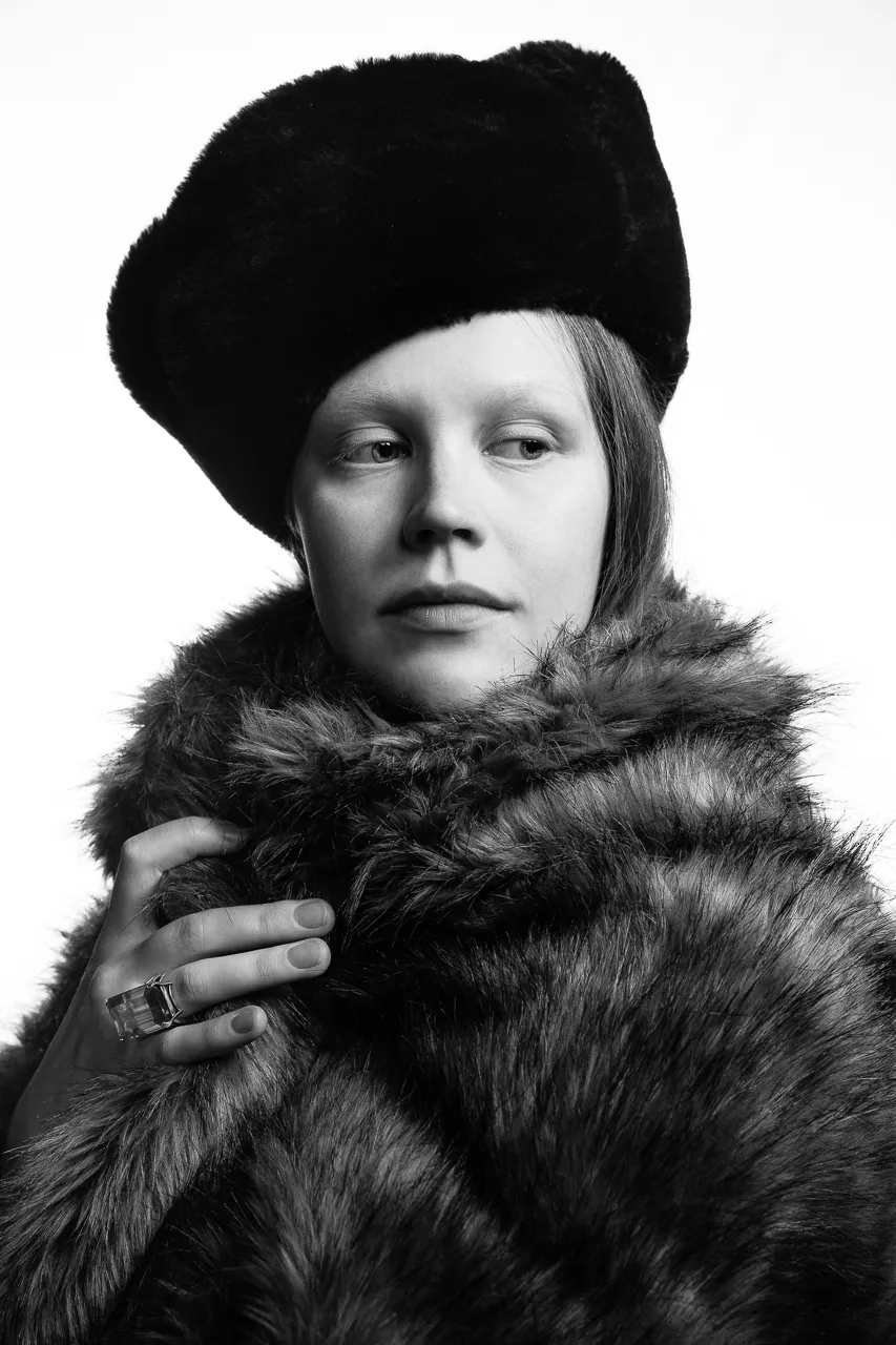 An actress in a fur hat and fur coat looks away, one ringed hand at her collar.