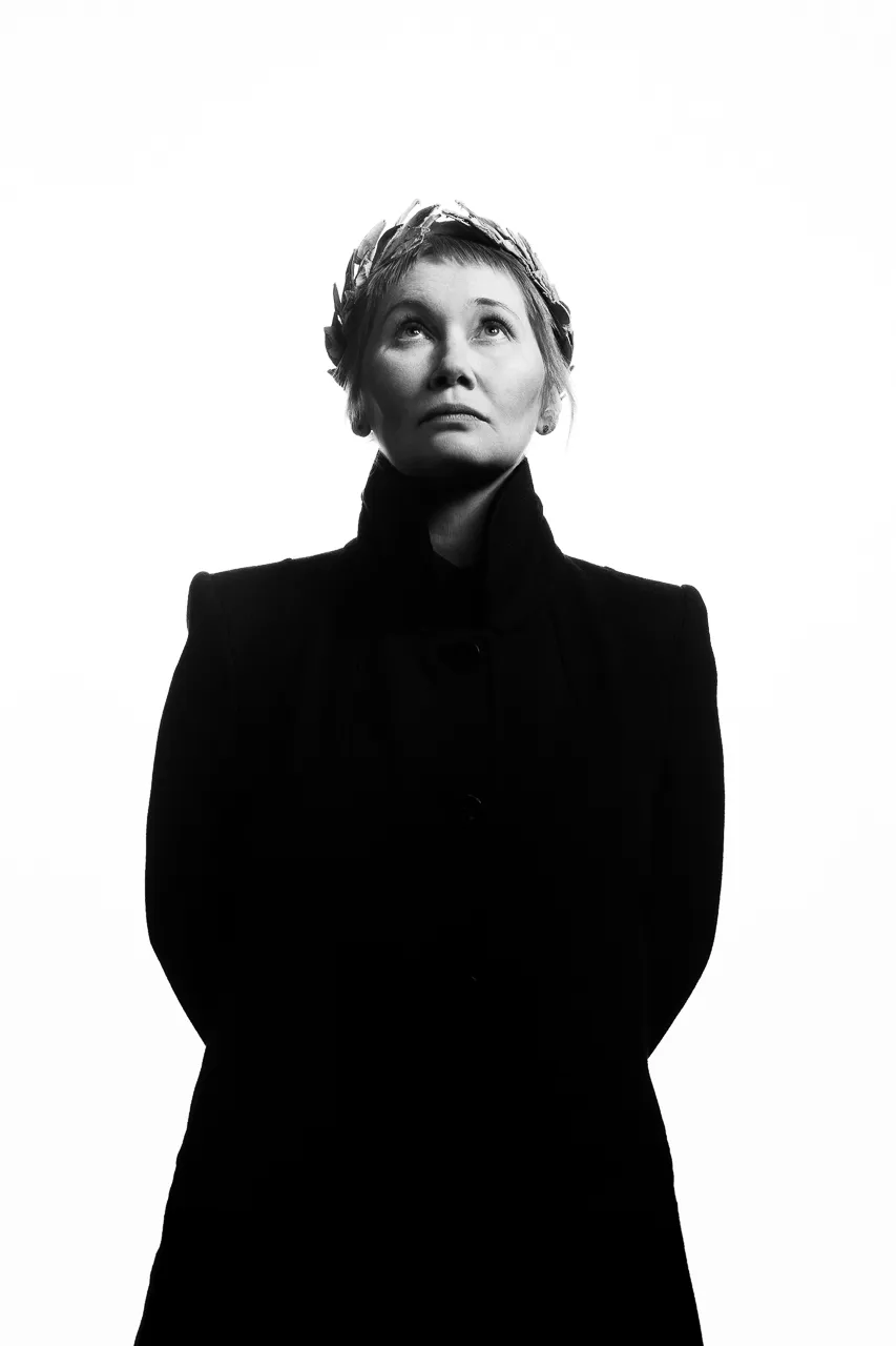An actress in a black coat looks upward, her hands behind her back.