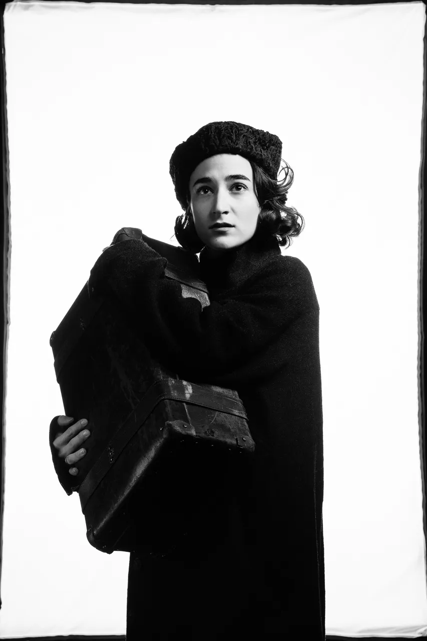 A young actress in a dark cap hugs a battered old suitcase.