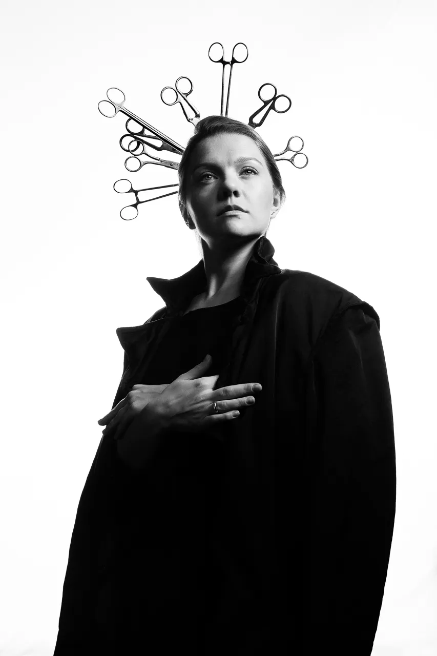 An actress wears a halo of surgical scissors, one hand on her chest.