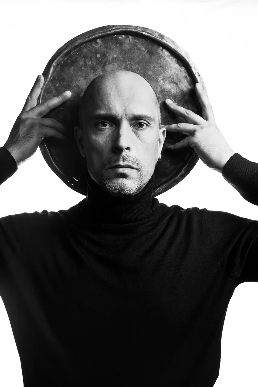 A bald actor holds an old metal basin behind his head like a halo.