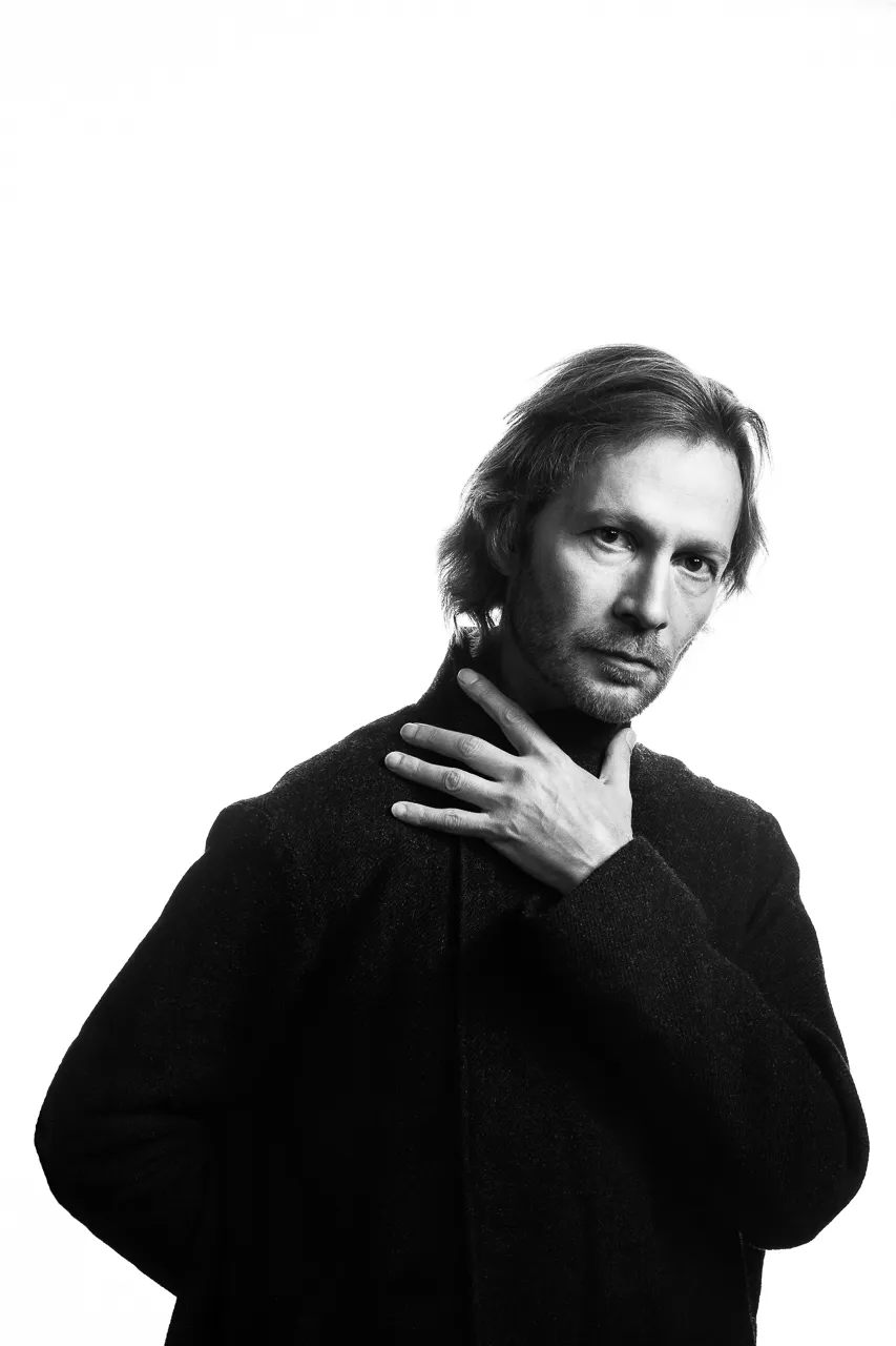 An actor with shoulder-length hair holds a hand to his throat.
