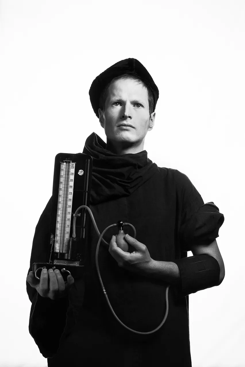 An actor in a black cap holds an old blood-pressure gauge and its bulb.
