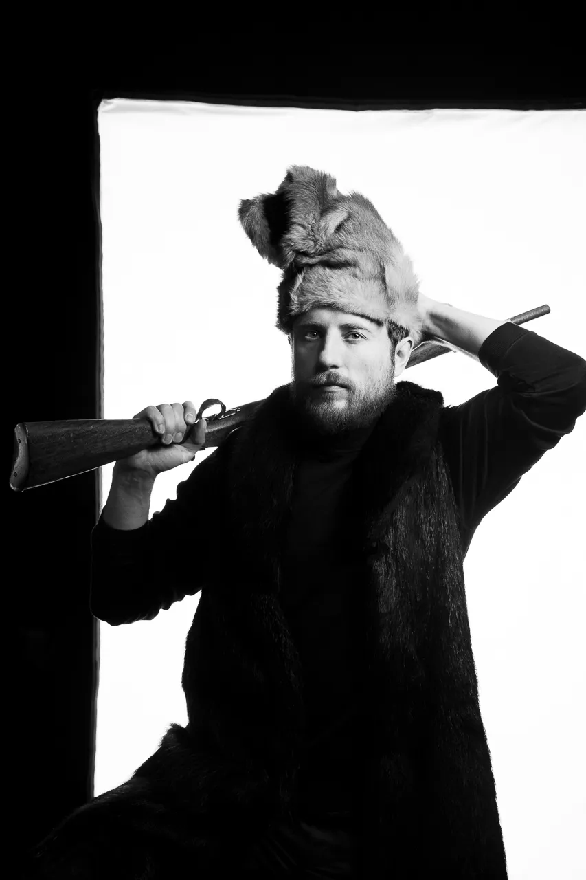 A bearded actor in a fur hat rests a rifle on his shoulder.