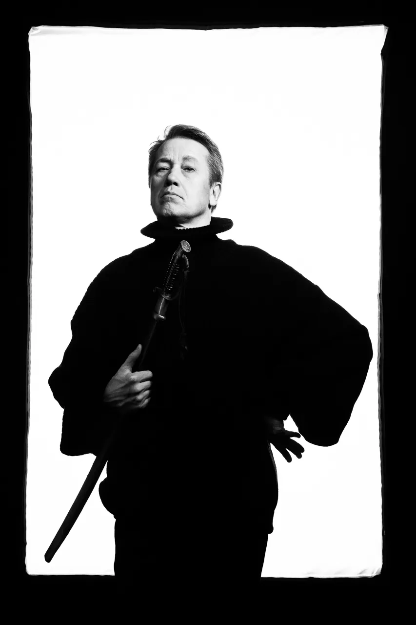 An actor in a wide black coat holds a sabre, one hand on his hip.