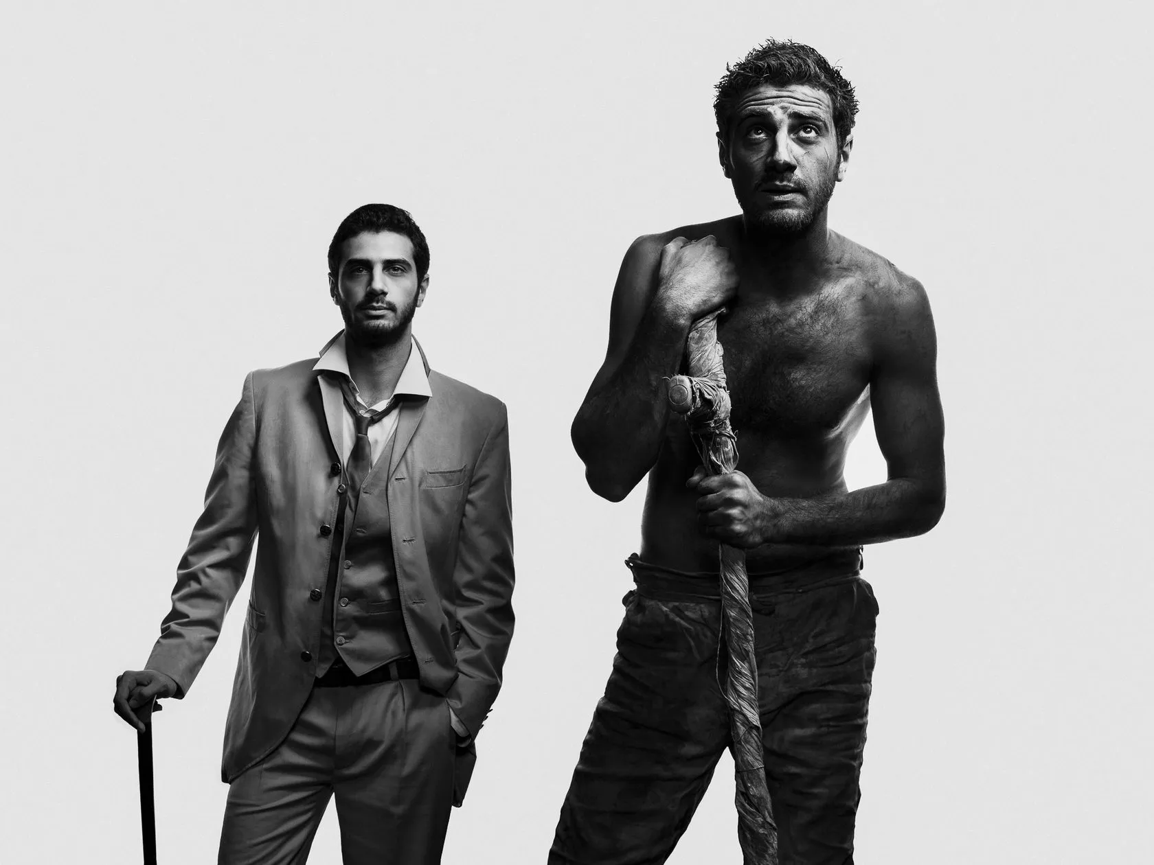 Two actors in costume: a man in a suit leaning on a cane, and a shirtless man gripping a rope-bound staff.