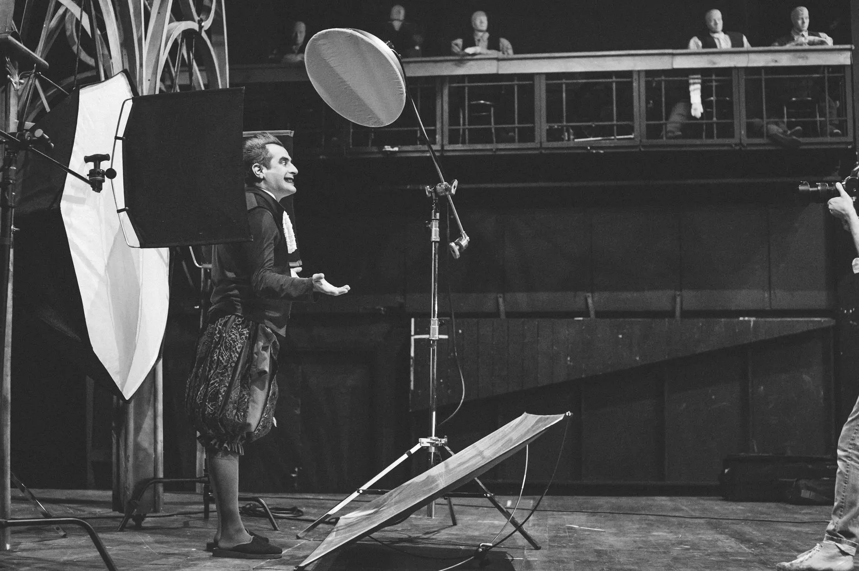 Behind the scenes: an actor in puffed breeches spreads his hands as people watch from the balcony.